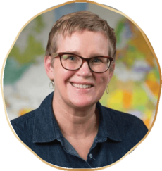 Melissa Paulsen Woman with short blond hair, wearing tortoiseshell glasses and a dark blue shirt, smiles in front of a colorful, blurred background.