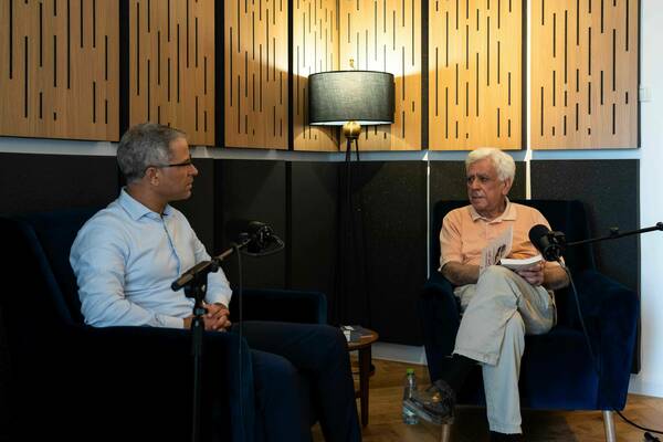 Two men sit in blue velvet chairs in a recording studio. A man with grey hair and glasses in a light blue shirt listens to a white-haired man in a peach polo holding an open book. Microphones are visible between them.