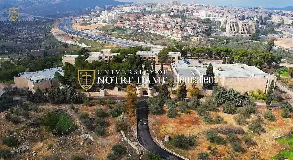 Aerial view of the University of Notre Dame at Tantur Ecumenical Institute in Jerusalem, with beige stone buildings, green trees, a winding road, and a city in the distance.