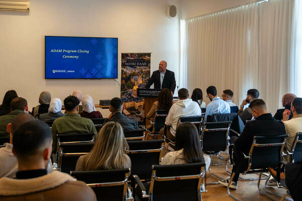 A bald man in a dark suit speaks from a podium to an audience at the ADAM Program Closing Ceremony. A screen displays the event title, and a banner shows the University of Notre Dame at Tantur, Jerusalem.