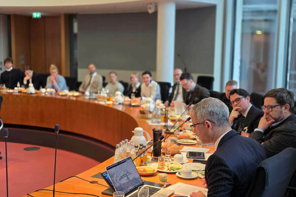 A man in a pinstripe suit reads documents at a curved conference table with several attentive attendees and laptops.