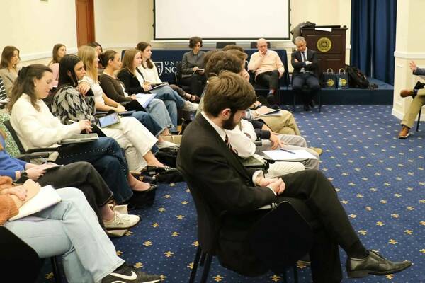 A speaker in a gray blazer addresses a diverse audience of students at a Notre Dame lecture, some taking notes.