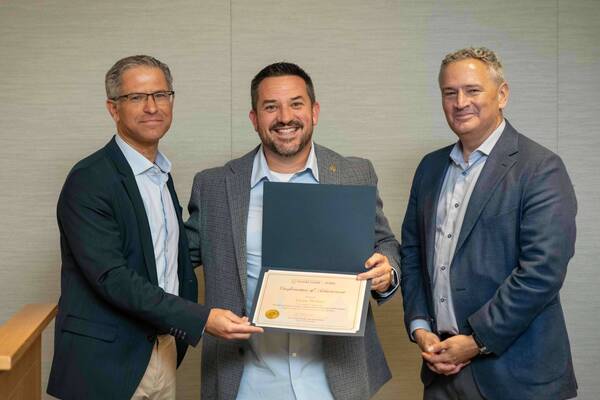 Three smiling men. Center, Jason Stoker receives a "Confirmation of Achievement" from Notre Dame Global from a man on his left.