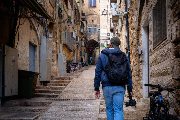 Person in blue jacket walks up a narrow, stone alley holding a camera. Old stone buildings with exposed wires & a Star of David.