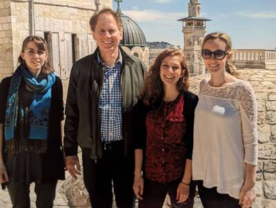 Four smiling people outdoors in front of stone buildings, a cross-topped dome, and a minaret-like tower.