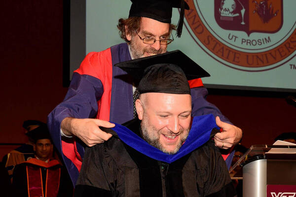 A man in purple and red academic regalia places a blue hood on a smiling man in a black gown at a Virginia Tech ceremony.