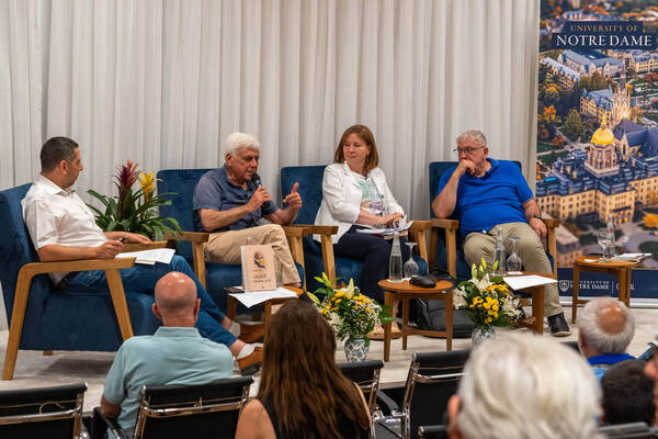 Four panelists on stage, one man speaking. A woman and two other men listen. Banner with University of Notre Dame Golden Dome.