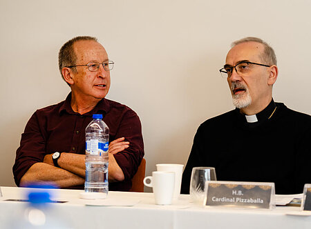 Cardinal Pizzaballa, in black clerical attire, speaks with a man in a burgundy shirt with crossed arms at a white table.