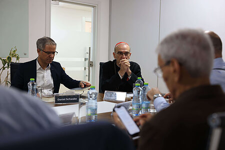 At a conference table, Daniel Schwake holds a notebook. A cardinal in a red zucchetto listens intently, hands clasped.