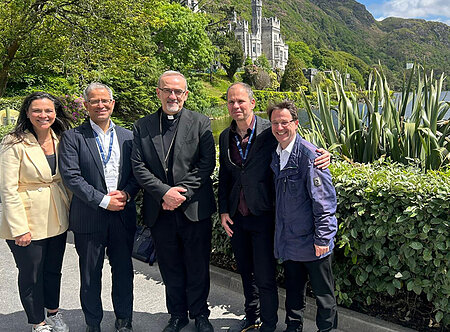 Five smiling individuals, including a man in black clerical attire, stand by Kylemore Abbey, a lake, and green hills.