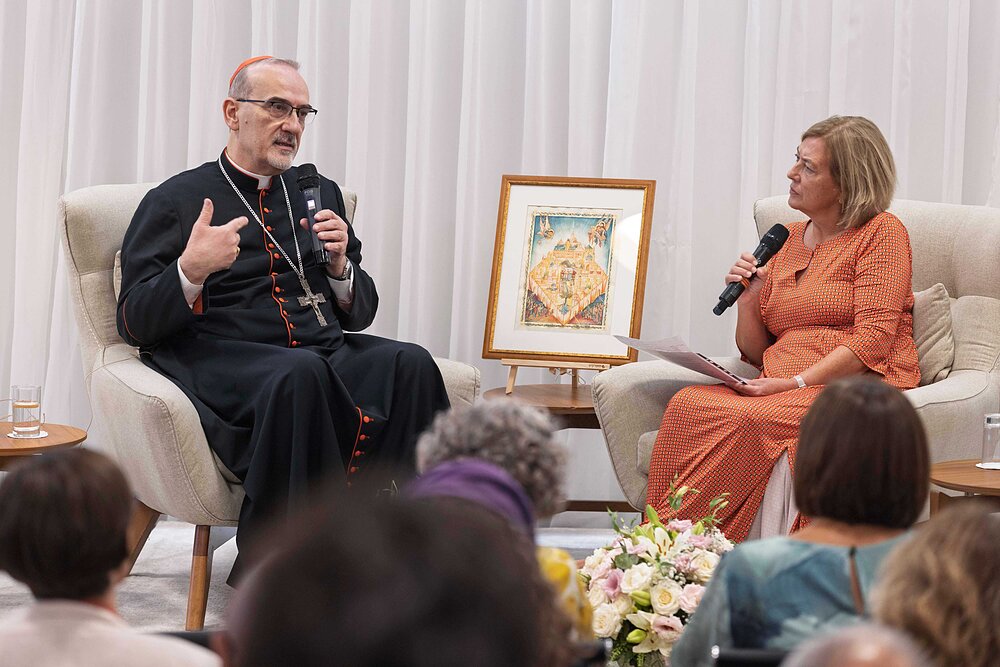 Cardinal wearing a cassock and cross speaks to a woman in an orange dress holding a microphone.